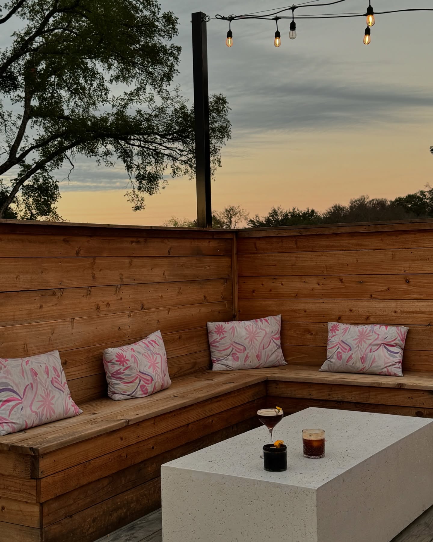 Outdoor patio at sunset with wooden benches, two drinks, and string lights.