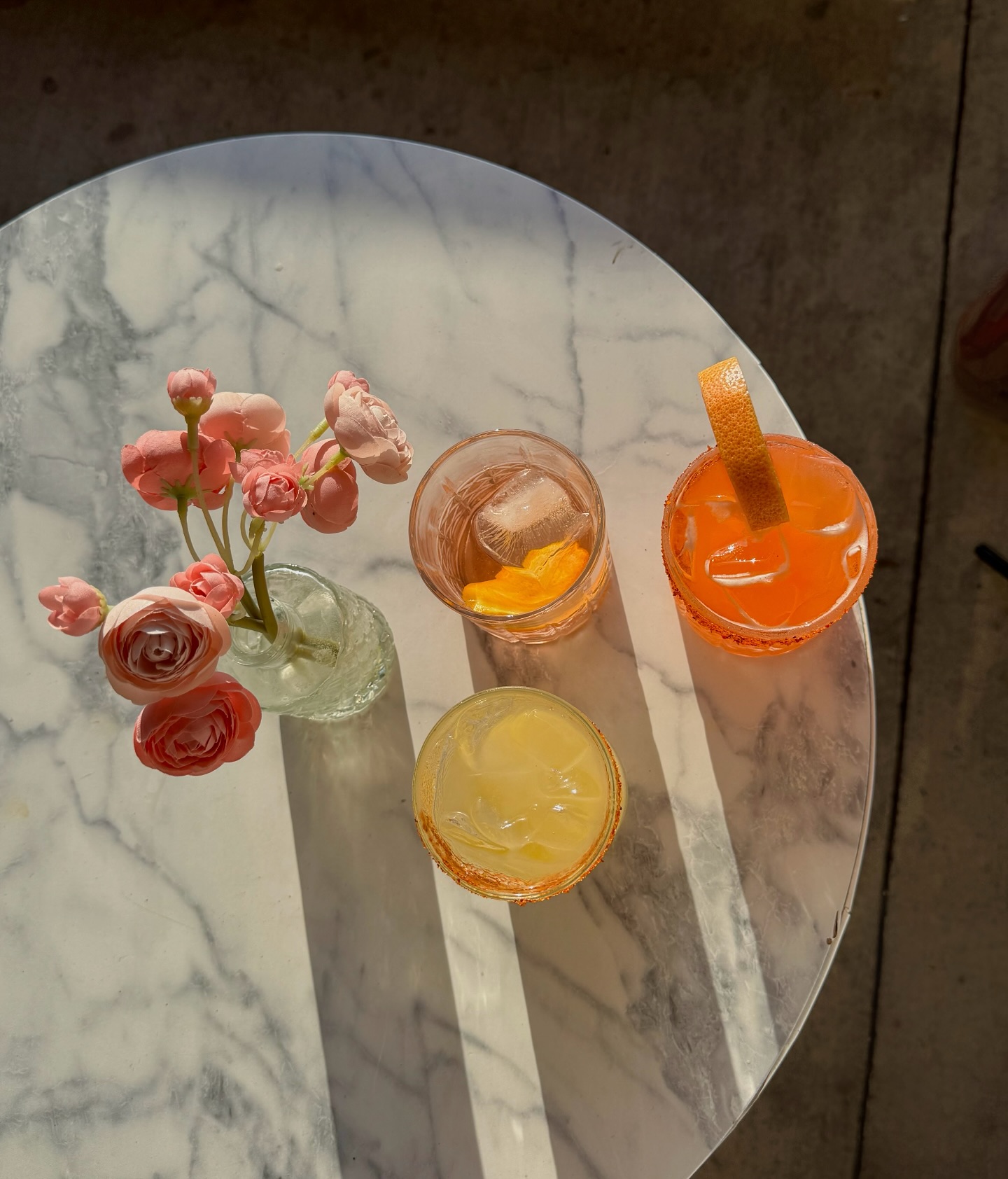 Pink flowers and three drinks on a marble table in sunlight.