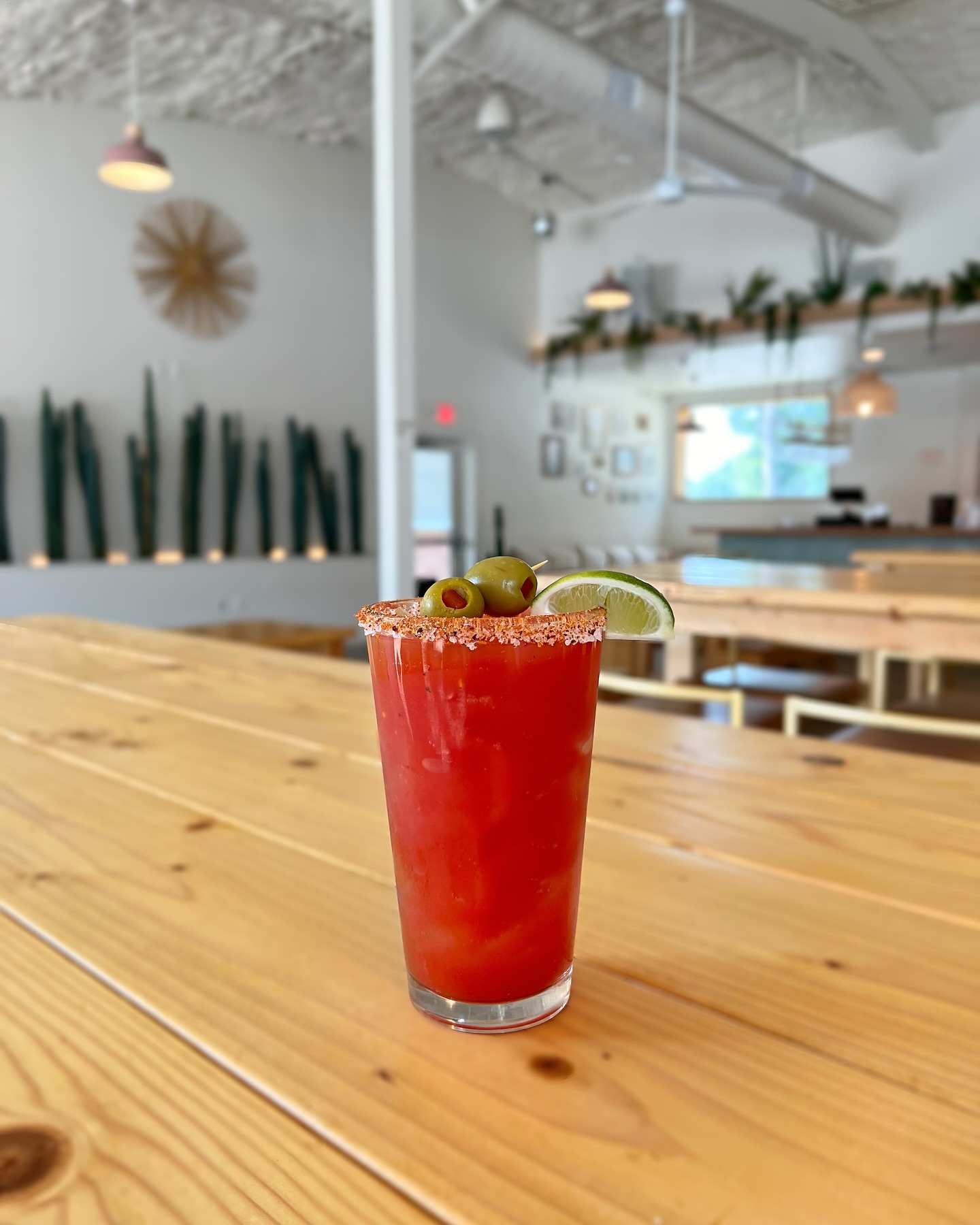 Red drink with spiced rim, olives, and lime on wooden table.
