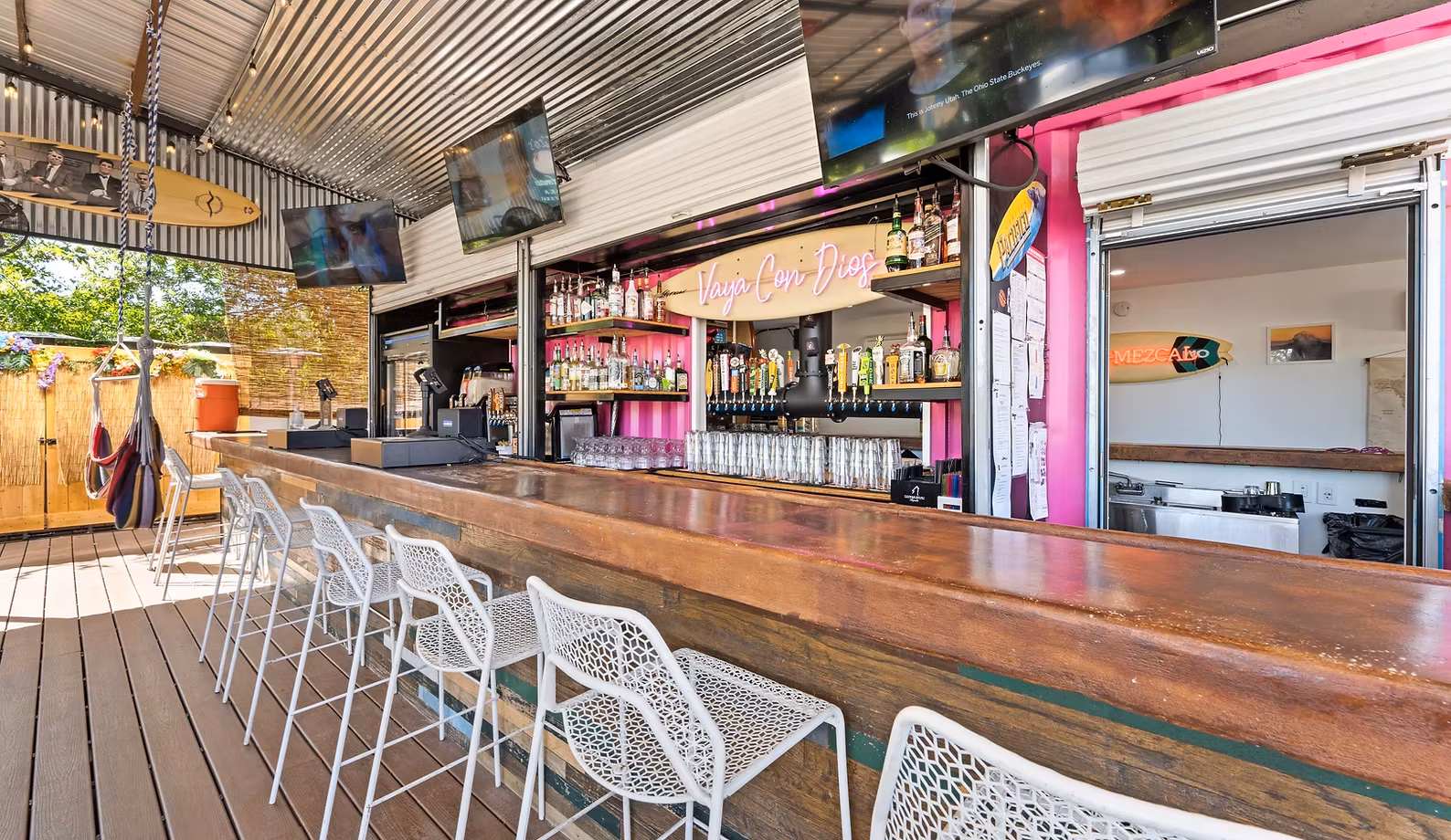 Outdoor bar with wooden counter, white stools, pink walls, and TVs.