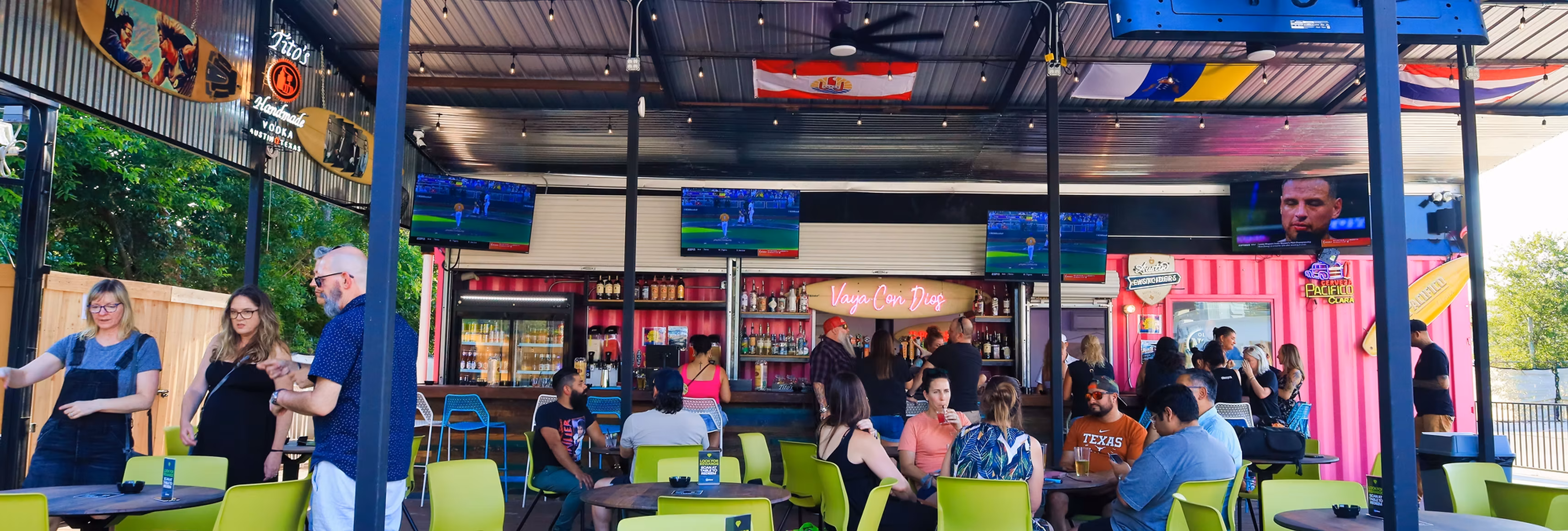 Crowded outdoor bar with sports on screens and pink container walls.