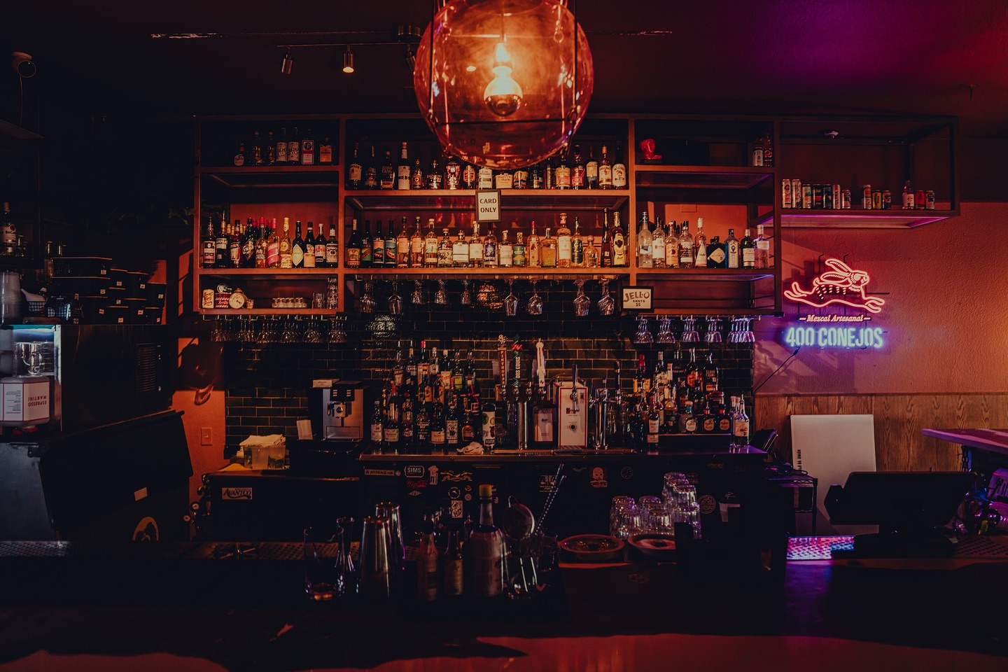 Dimly lit bar with liquor bottles on shelves and a neon rabbit sign.