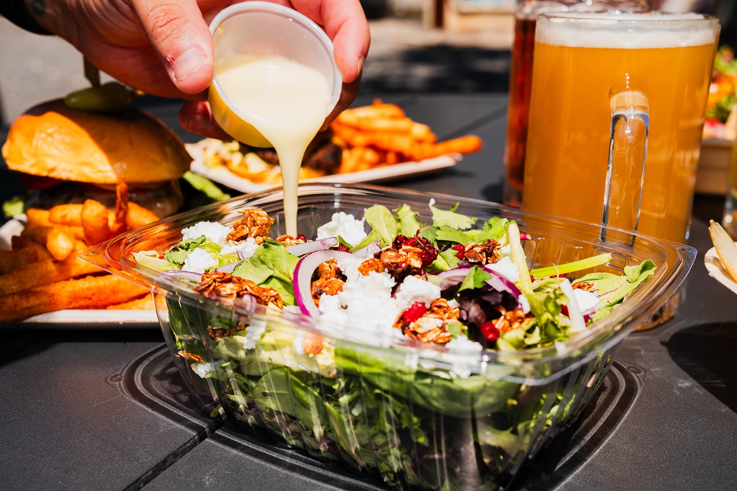 Hand pouring dressing onto a salad, with burger, fries, and beer.