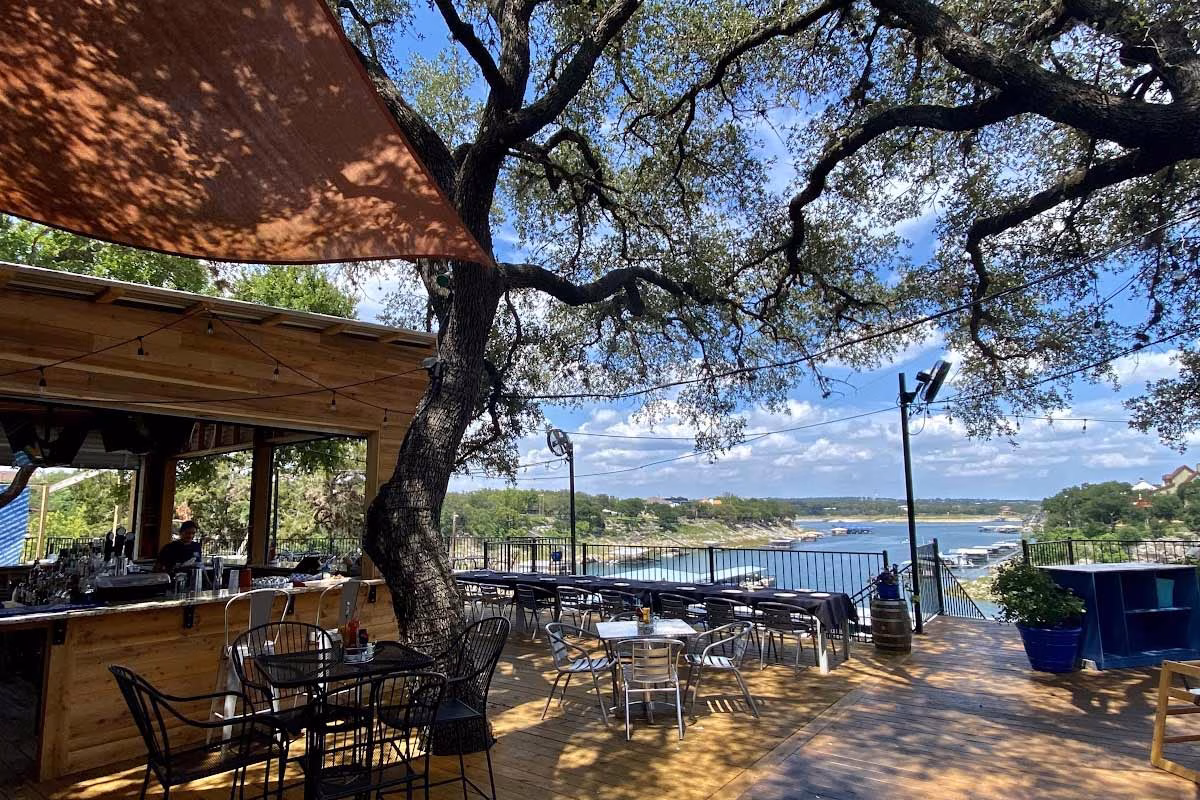 Lakeside restaurant patio with a bar, shaded by a large tree.