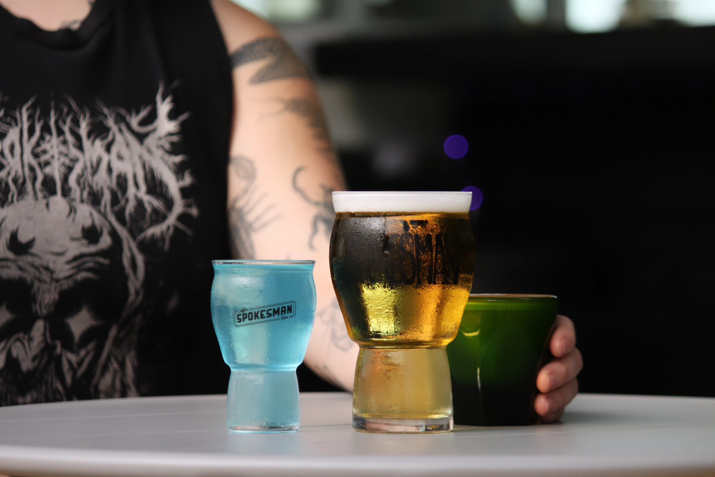 Tattooed person's arm near blue drink, cold beer, green glass.