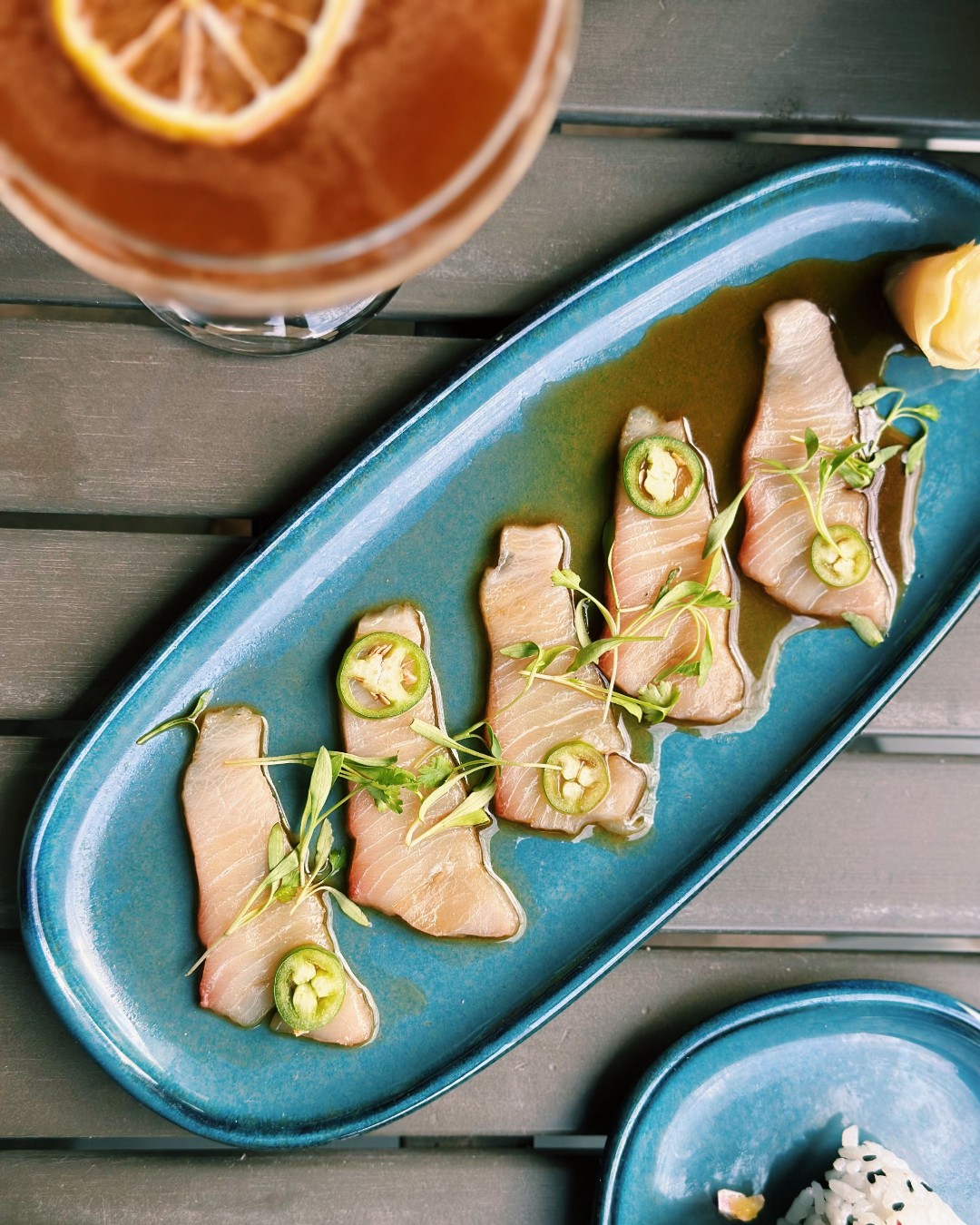 Platter of sliced fish with jalapenos and greens, drink with lemon.