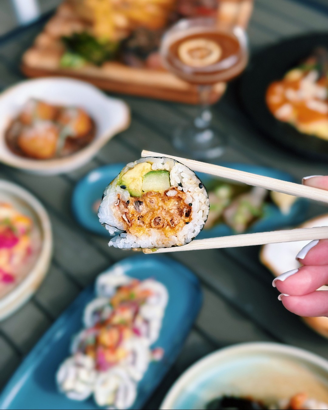 Chopsticks hold a sushi roll with avocado, cucumber, and crunchy filling.