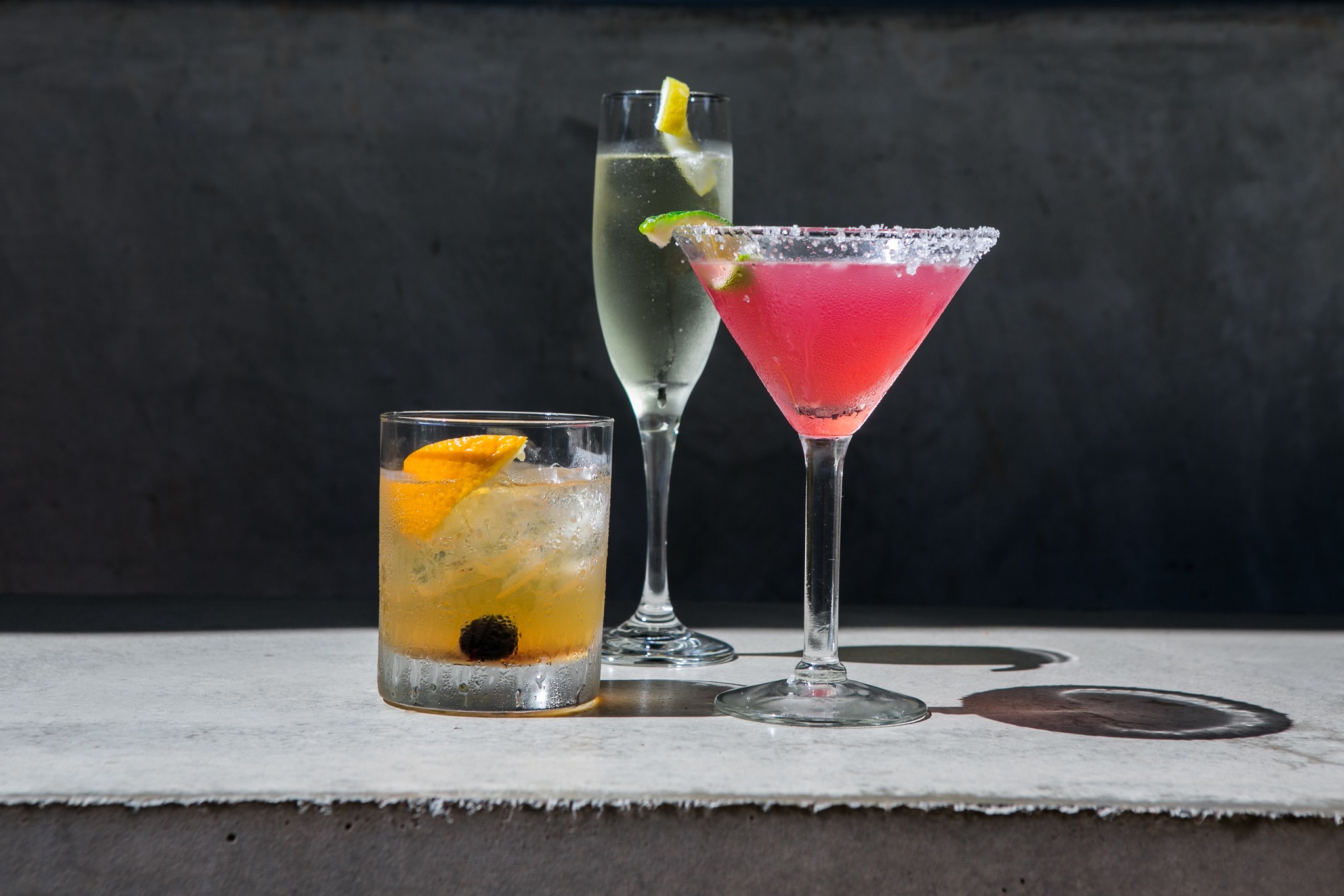 Three cocktails, garnished with fruit and salt, on a concrete surface.