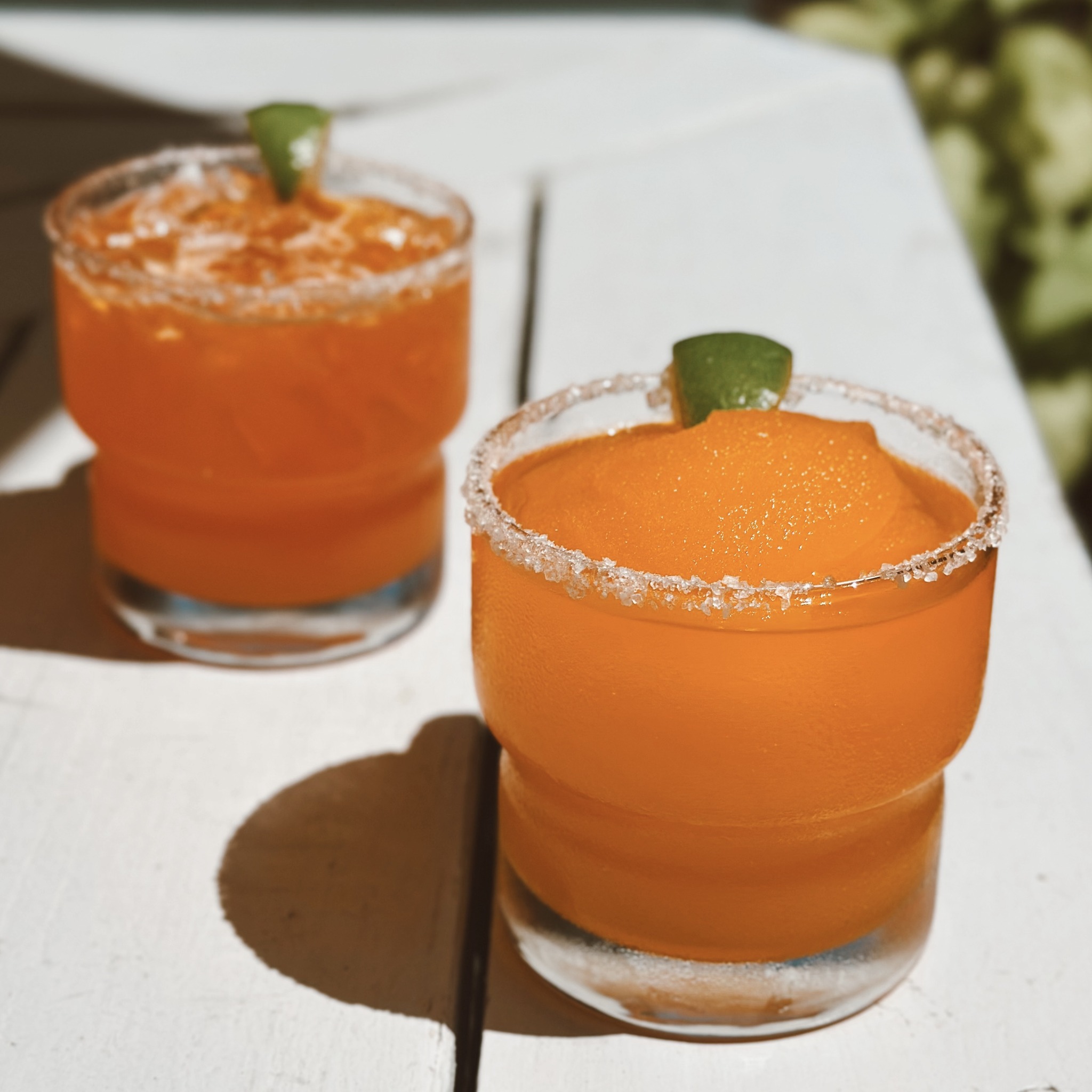 Two orange drinks with lime on white table, one frozen.