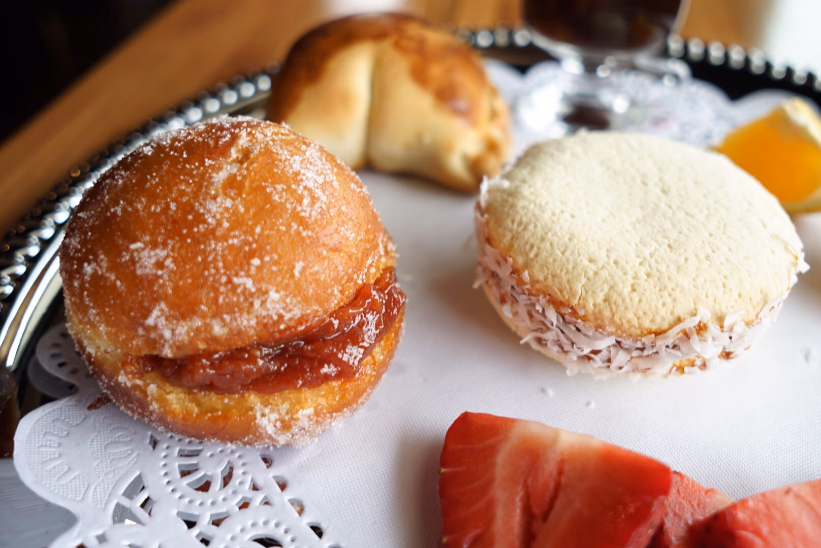 Sugar-dusted jam donut, coconut alfajor, and cut strawberries on a platter.