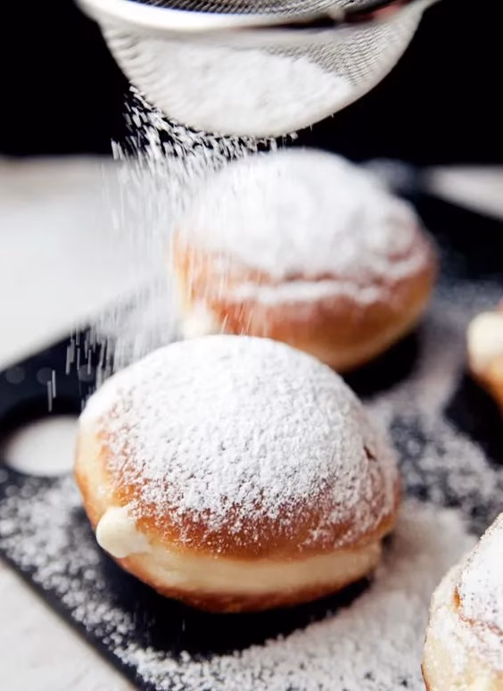 Powdered sugar being sifted onto cream-filled pastries on a dark surface.