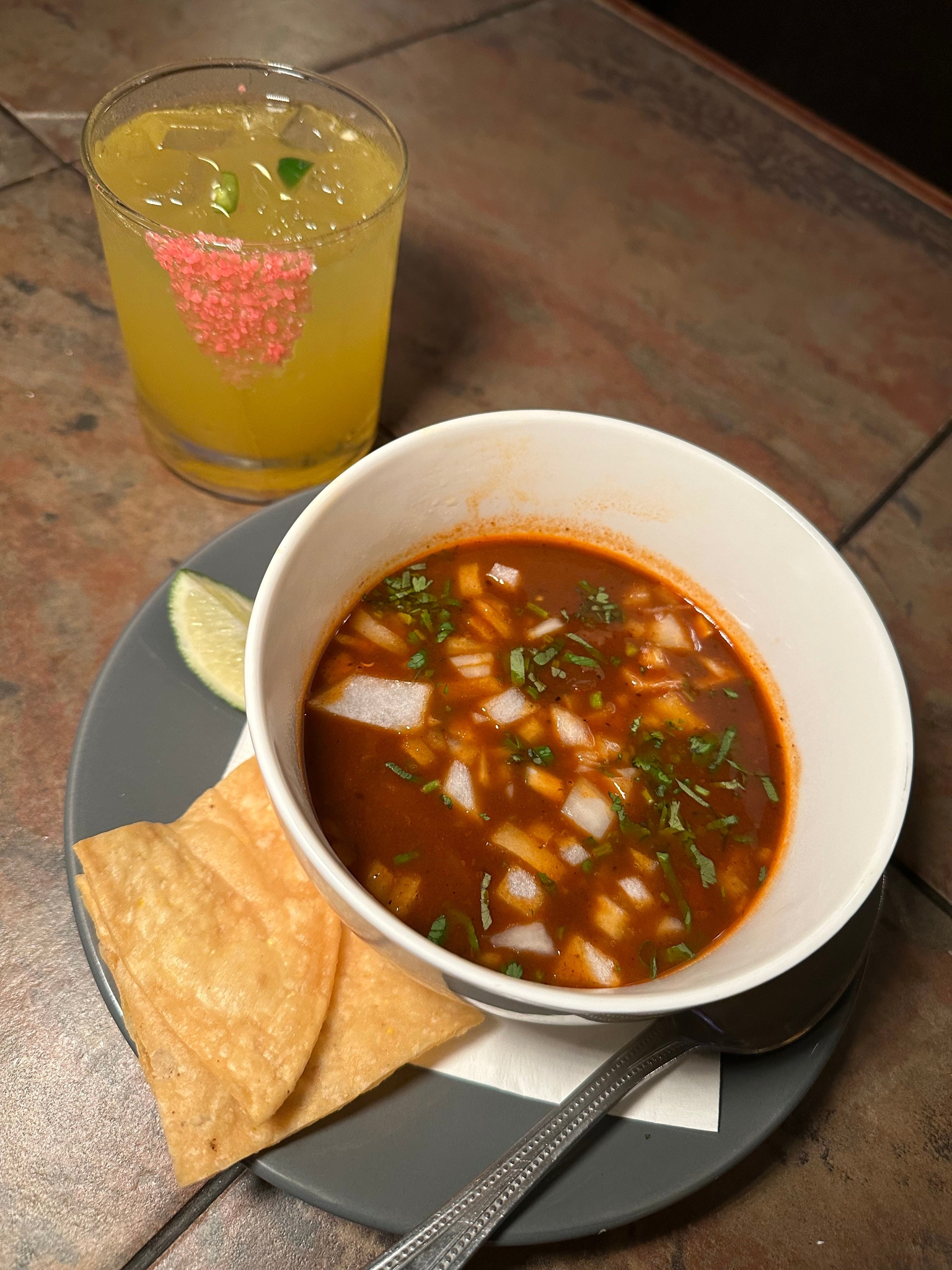 Reddish soup, tortilla chips, lime wedge, and a yellow drink with sprinkles.