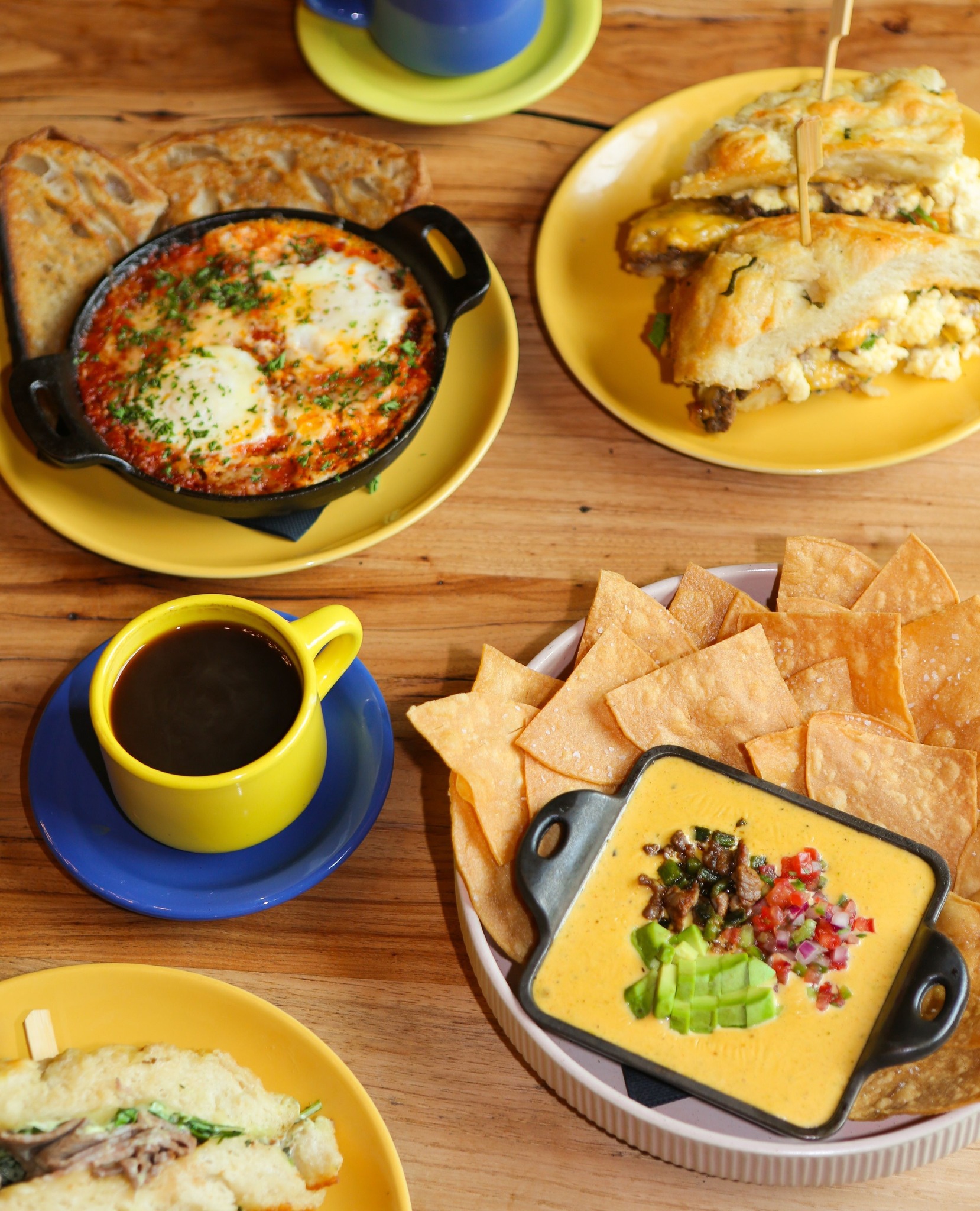 Breakfast spread with shakshuka, sandwiches, queso, chips, and coffee.