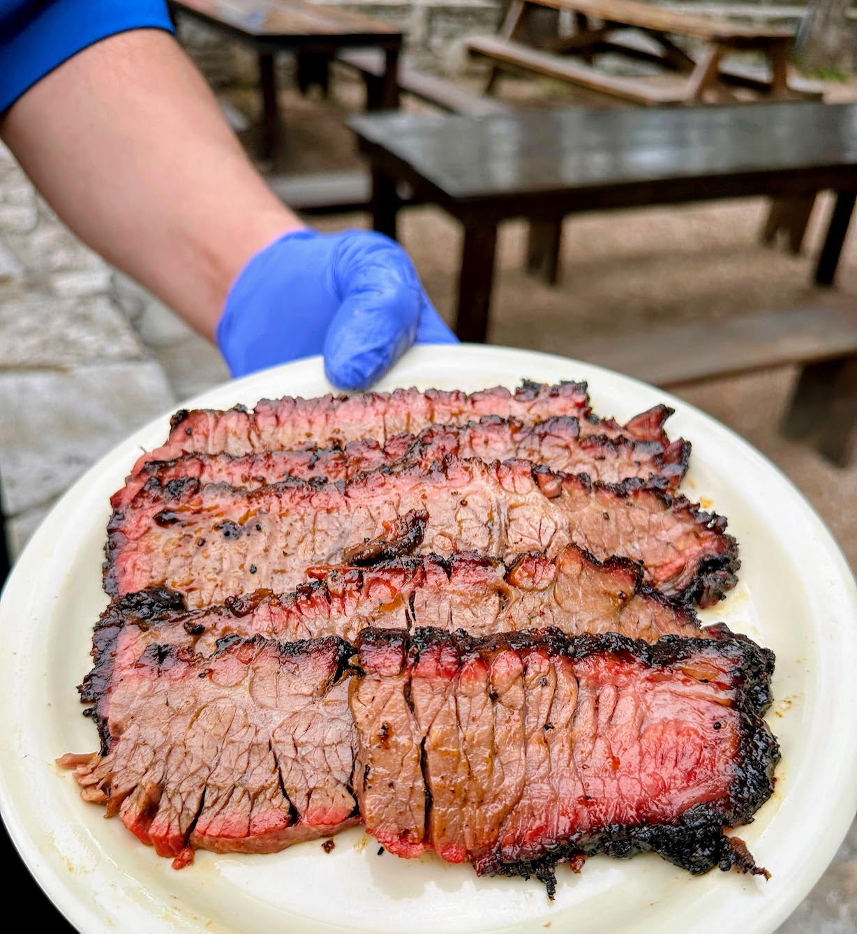 A gloved hand holds a plate of sliced, smoked brisket.
