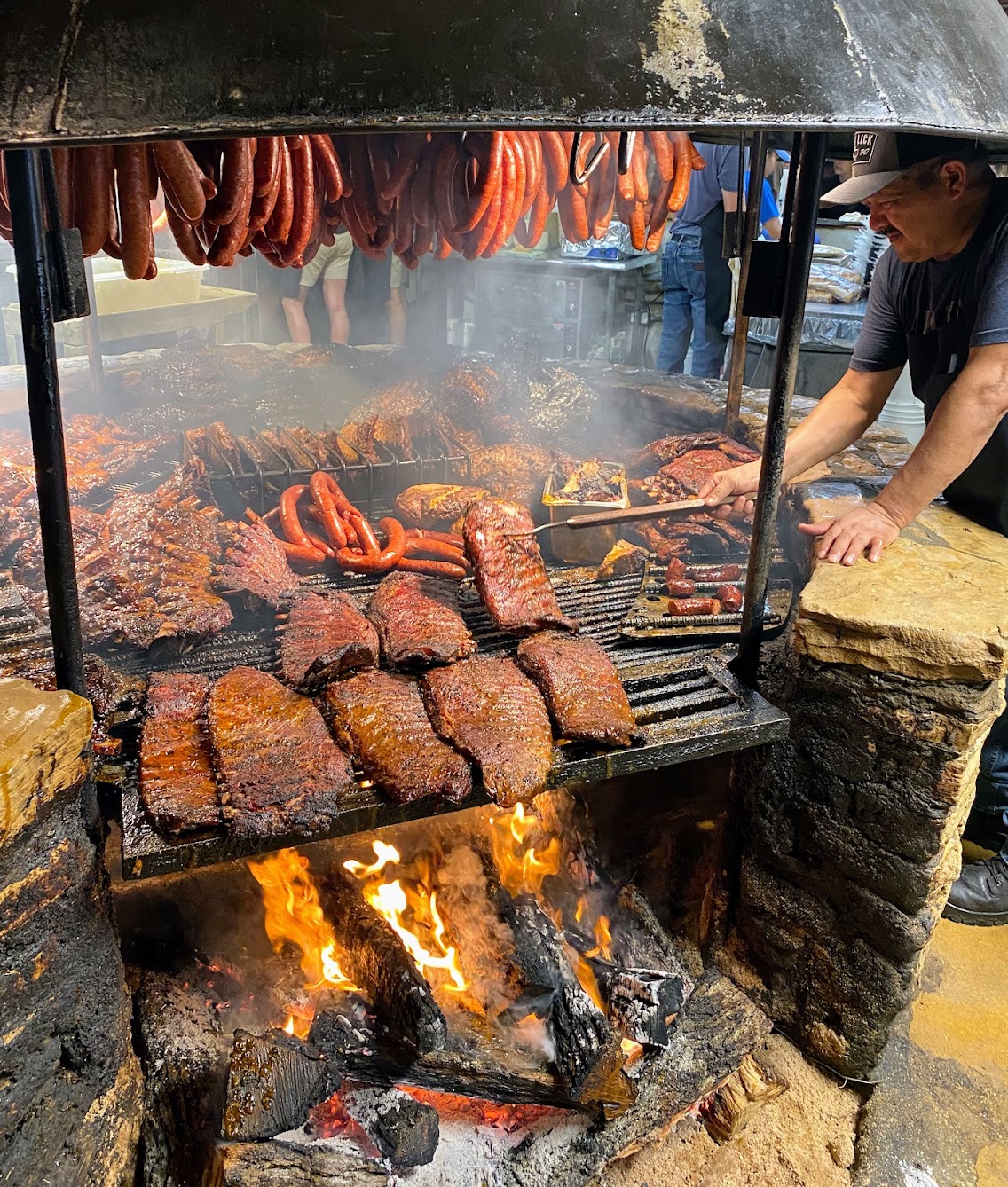 Man tending large smoky barbecue pit with hanging sausages and ribs.