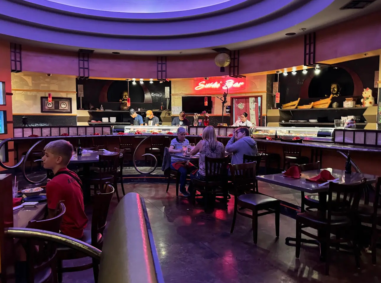 Interior of a sushi restaurant with patrons and a neon "Sushi Bar" sign.