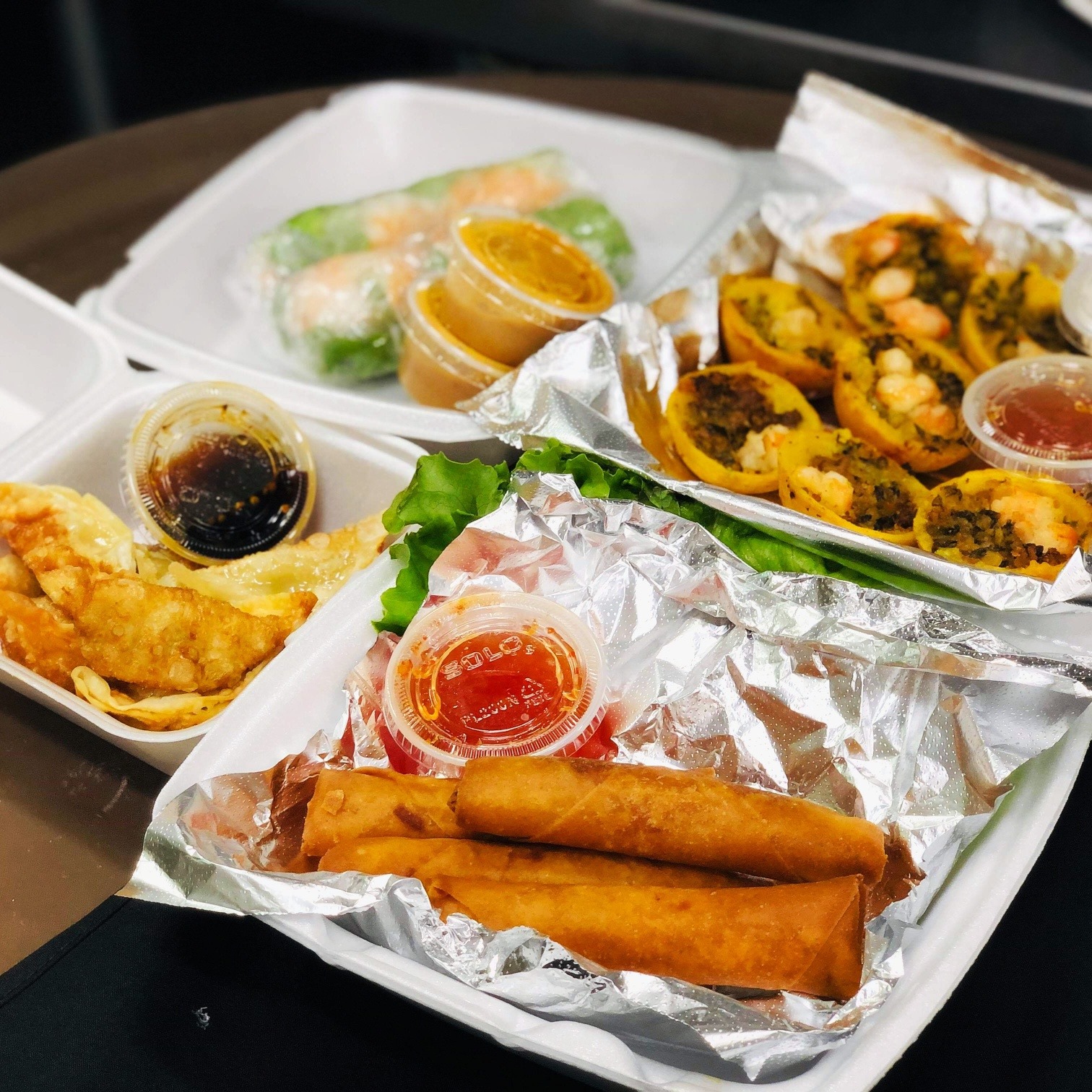 Takeout containers with fried spring rolls, dumplings, fresh rolls, and pancakes.
