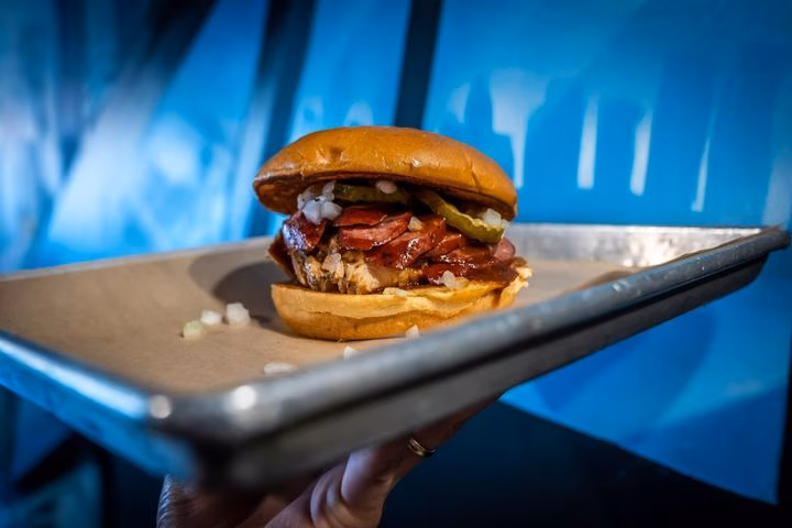 Sandwich with pulled meat, sausage, pickles, onions on a serving tray.