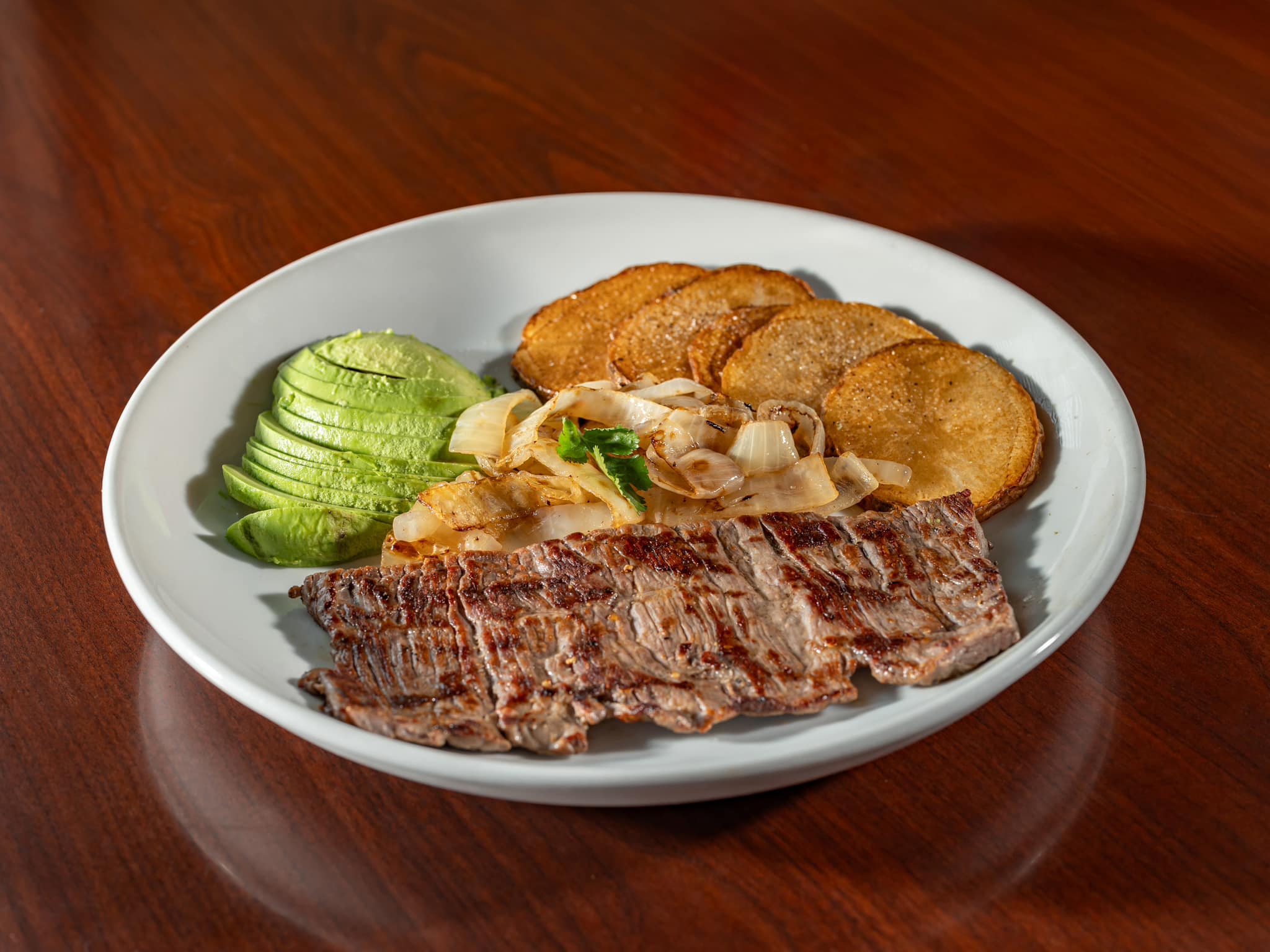 Grilled steak, avocado, onions, and potatoes on a white plate.