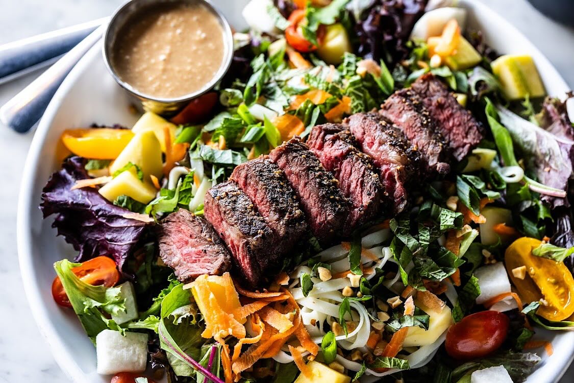 Sliced steak over a colorful salad with noodles and dressing.