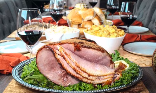 Carved ham on a platter, wine, and side dishes on a table.