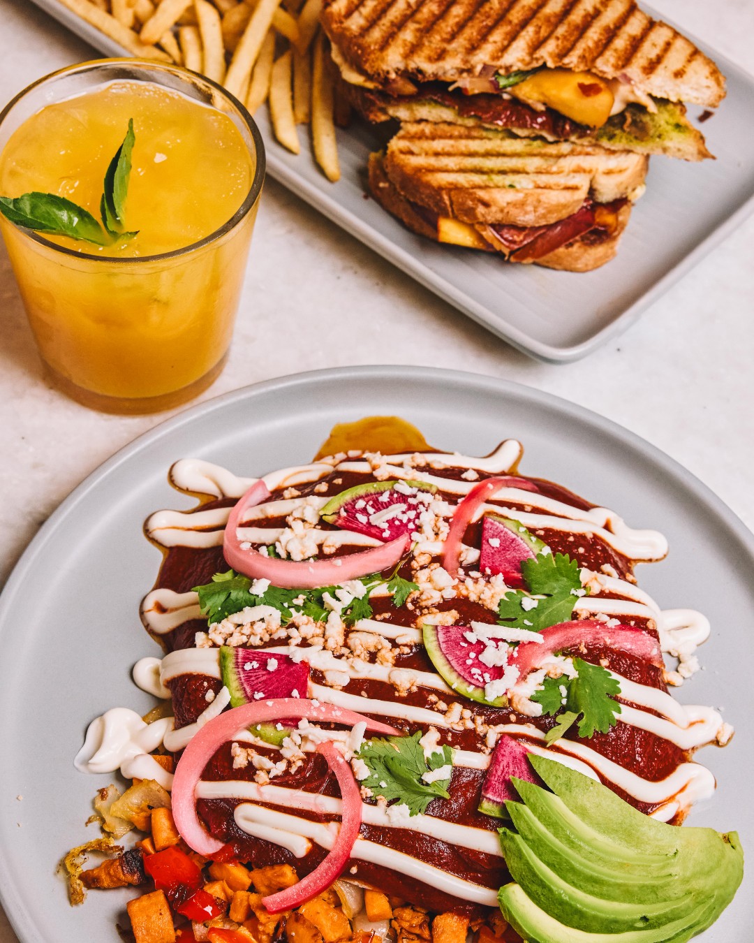 Plates of sauced dish, grilled sandwich, fries, and a yellow drink.