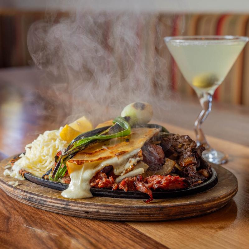 Sizzling platter with melted cheese, meats, vegetables, and a cocktail.