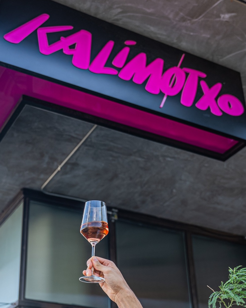 Hand holding rosé wine glass under a "KALIMOTXO" sign.