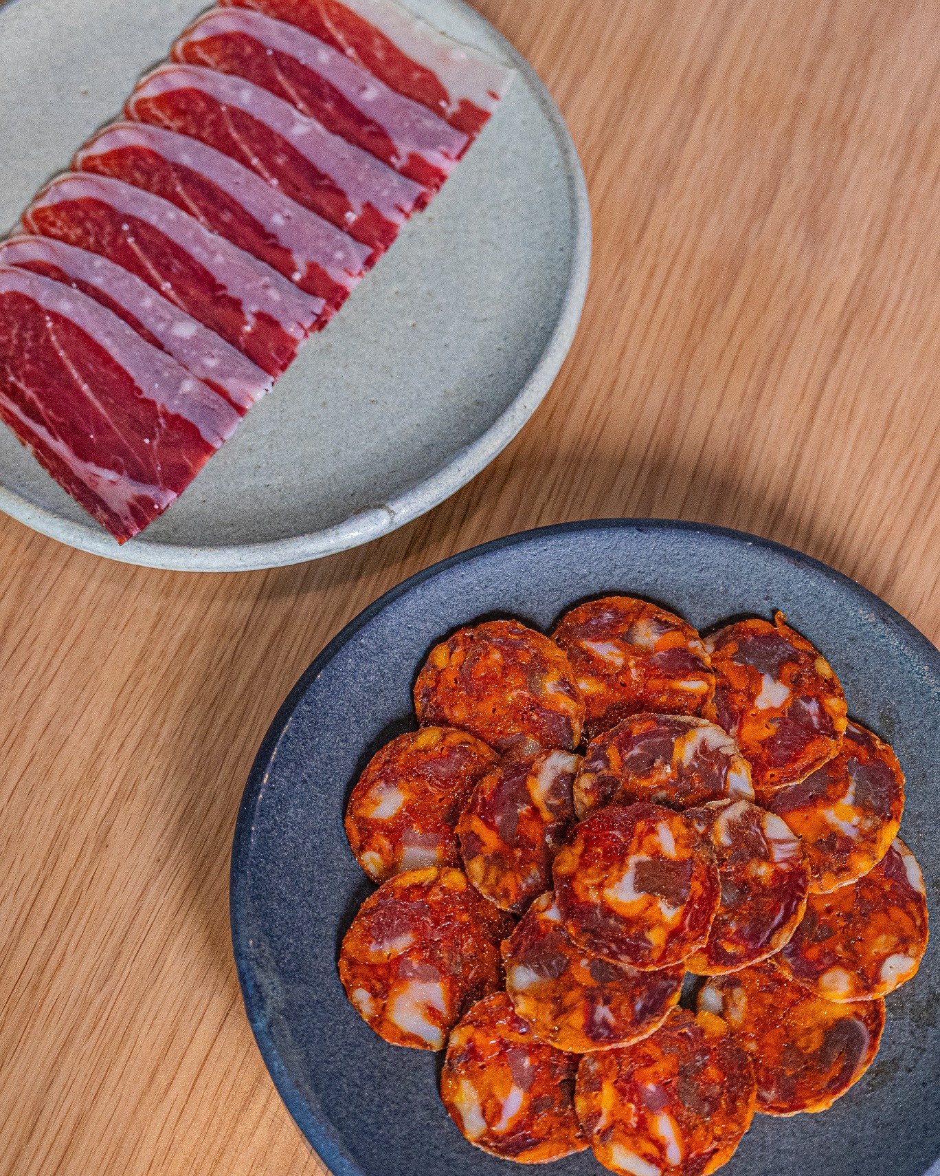 Two plates of sliced ham and chorizo on a wooden table.