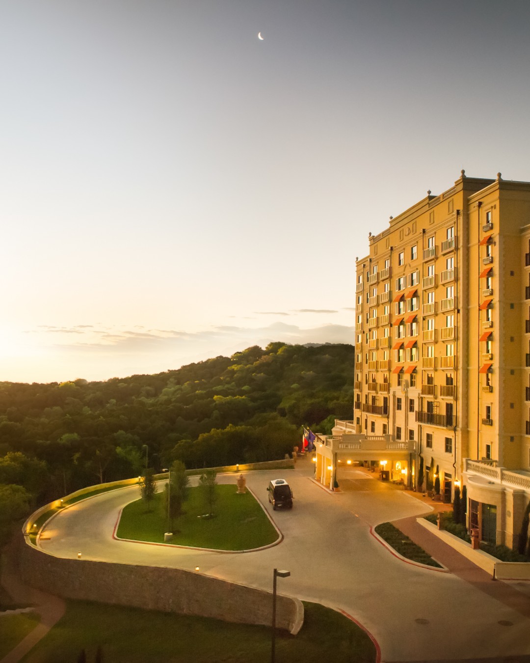 Hotel building, curved driveway, wooded hills, and crescent moon at dawn.
