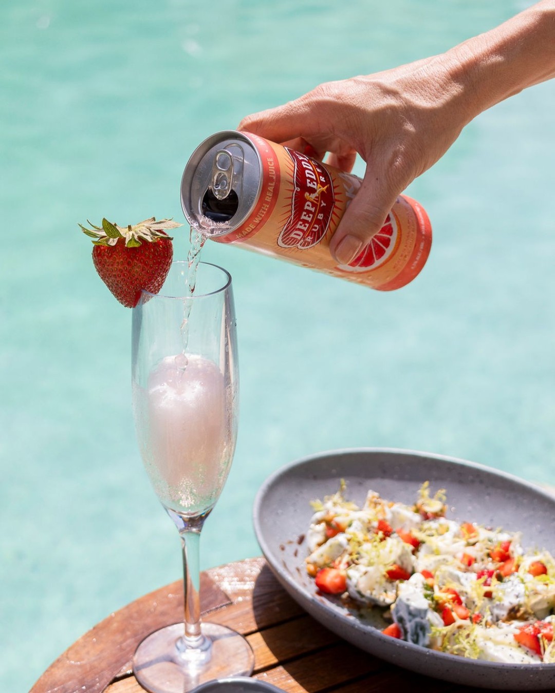 Hand pouring Deep Eddy Ruby Red from can into strawberry-garnished flute.