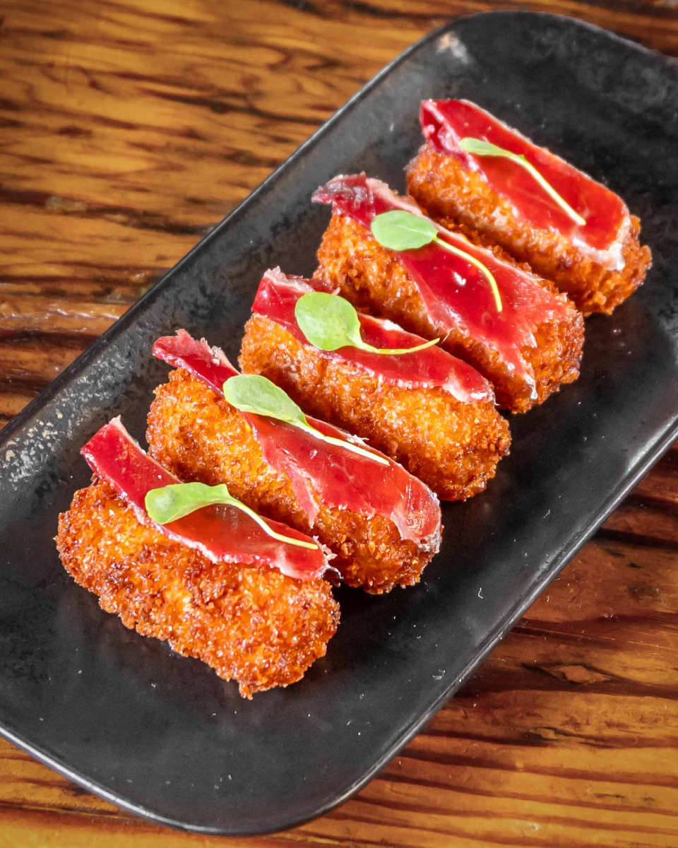 Five fried croquettes topped with cured ham and green garnish on black plate.