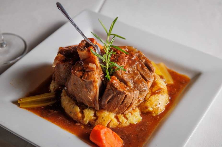 Braised meat shank with polenta, sauce, rosemary, and fork on plate.
