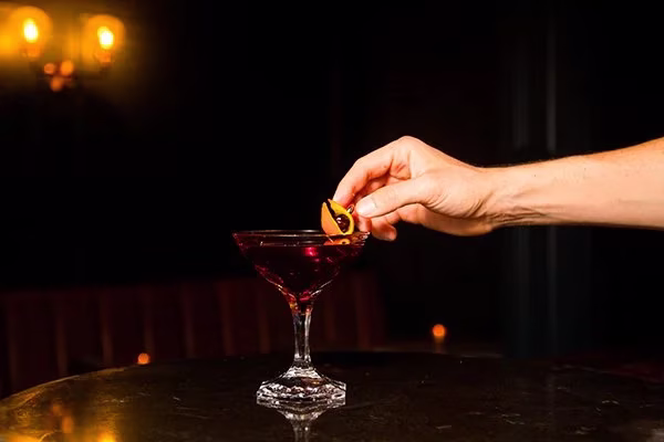 Hand garnishing red cocktail with a twisted orange peel.