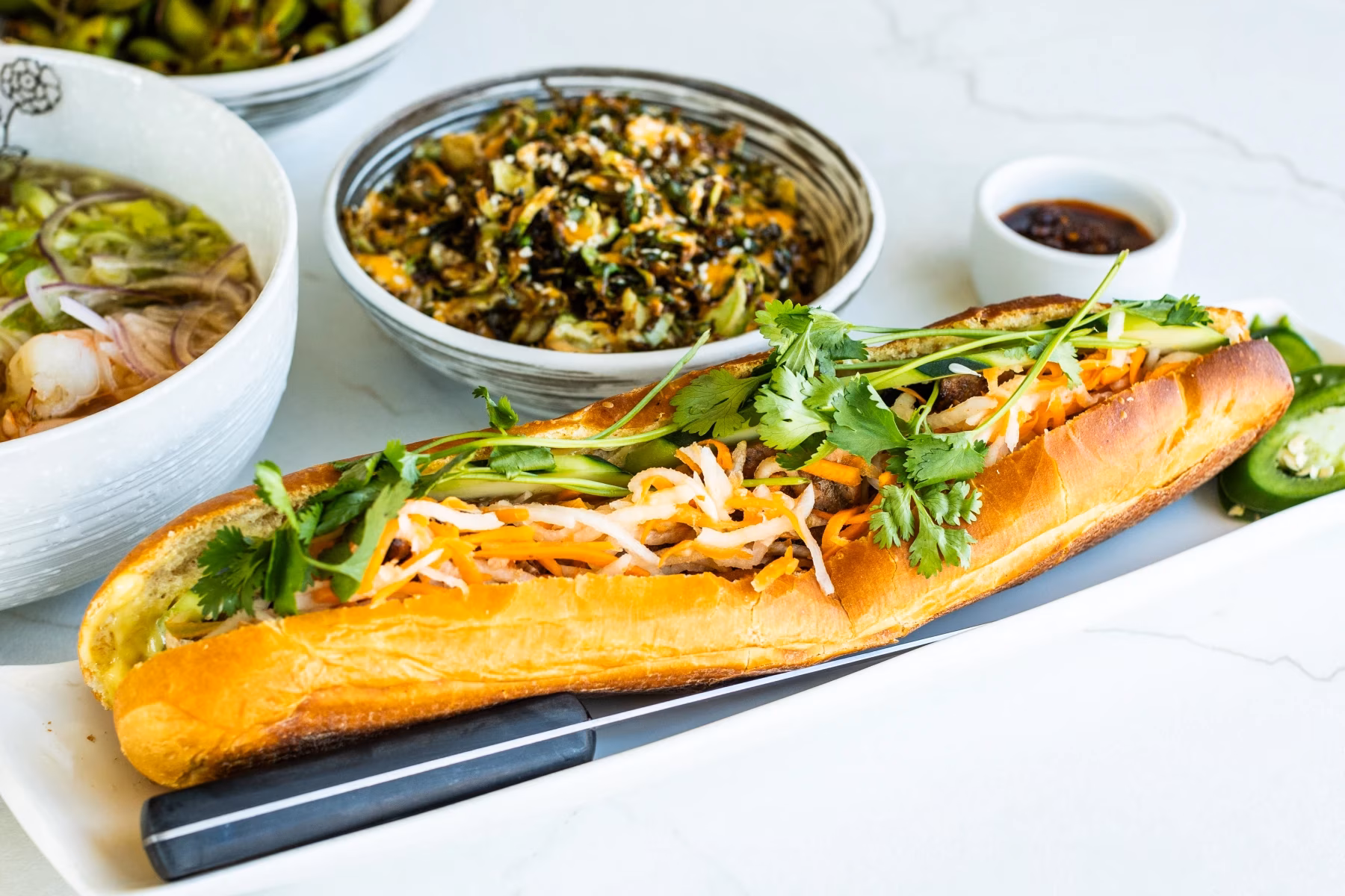 Vietnamese banh mi sandwich, pho, and side dishes on a white table.