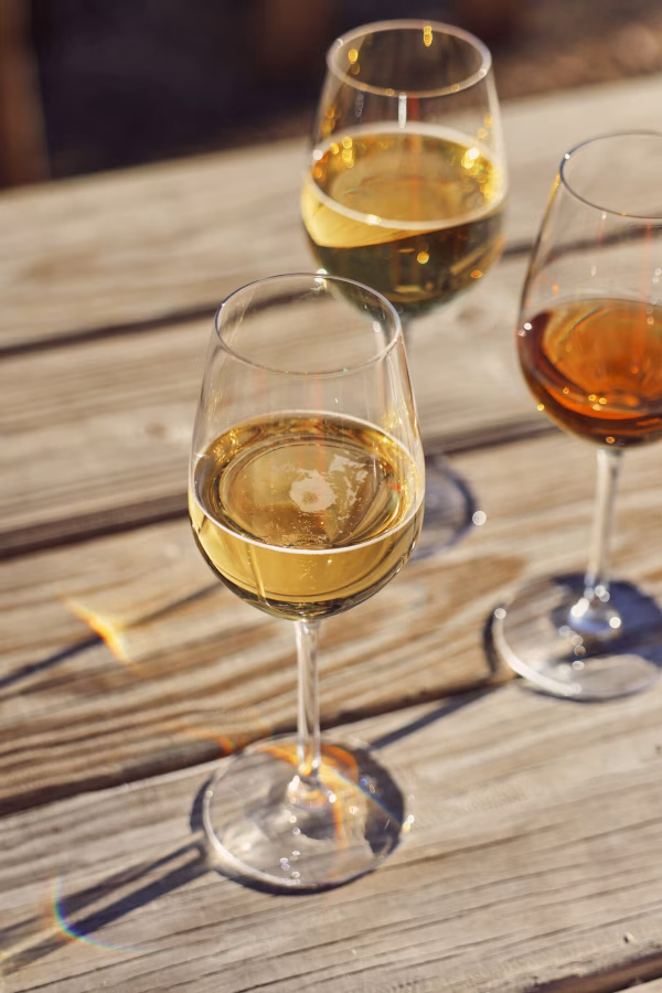 Three glasses of white and amber wine on a sunlit wooden table.