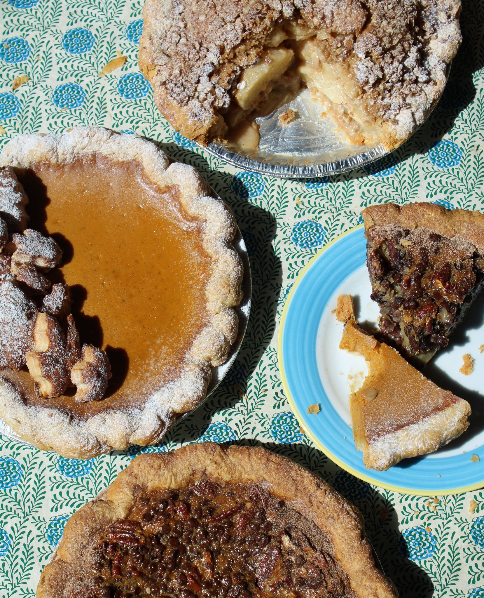 Apple, pumpkin, and pecan pies, some with slices, on a tablecloth.