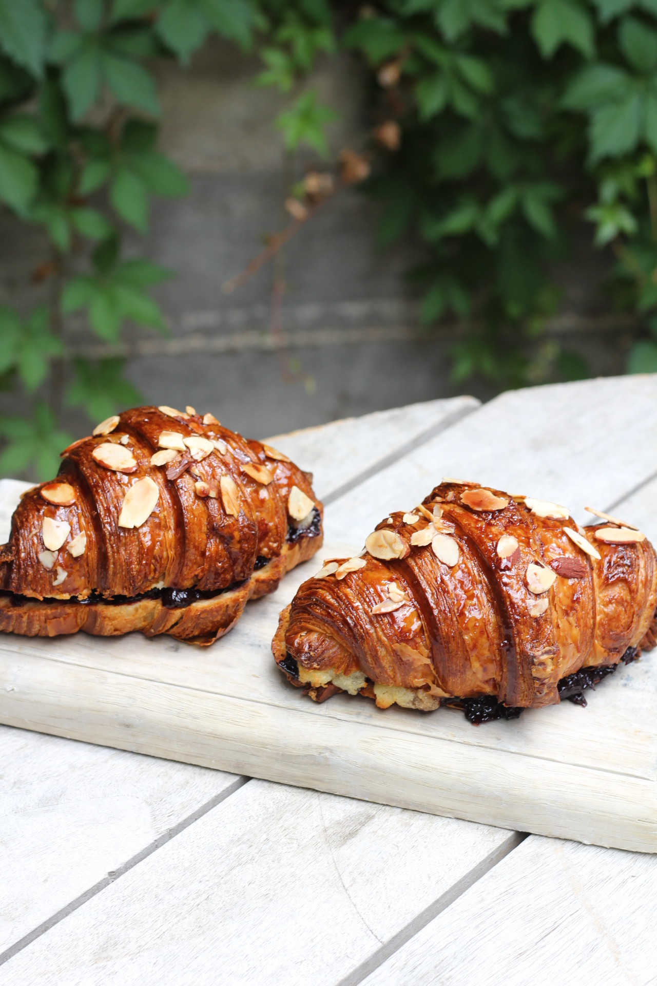 Two glazed almond croissants with dark filling on a light wooden board.