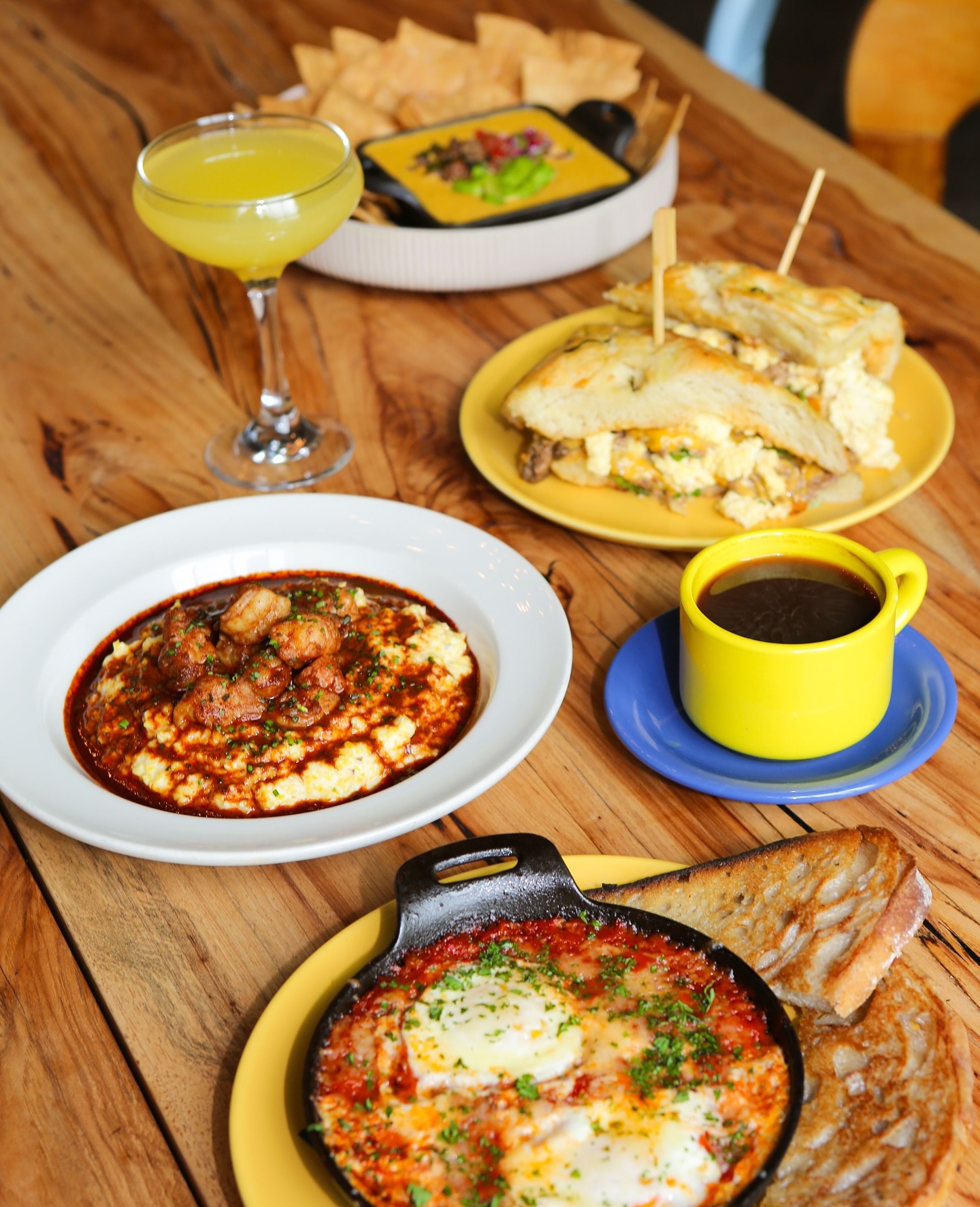 Shrimp and grits, baked eggs, sandwiches, and drinks on a wooden table.