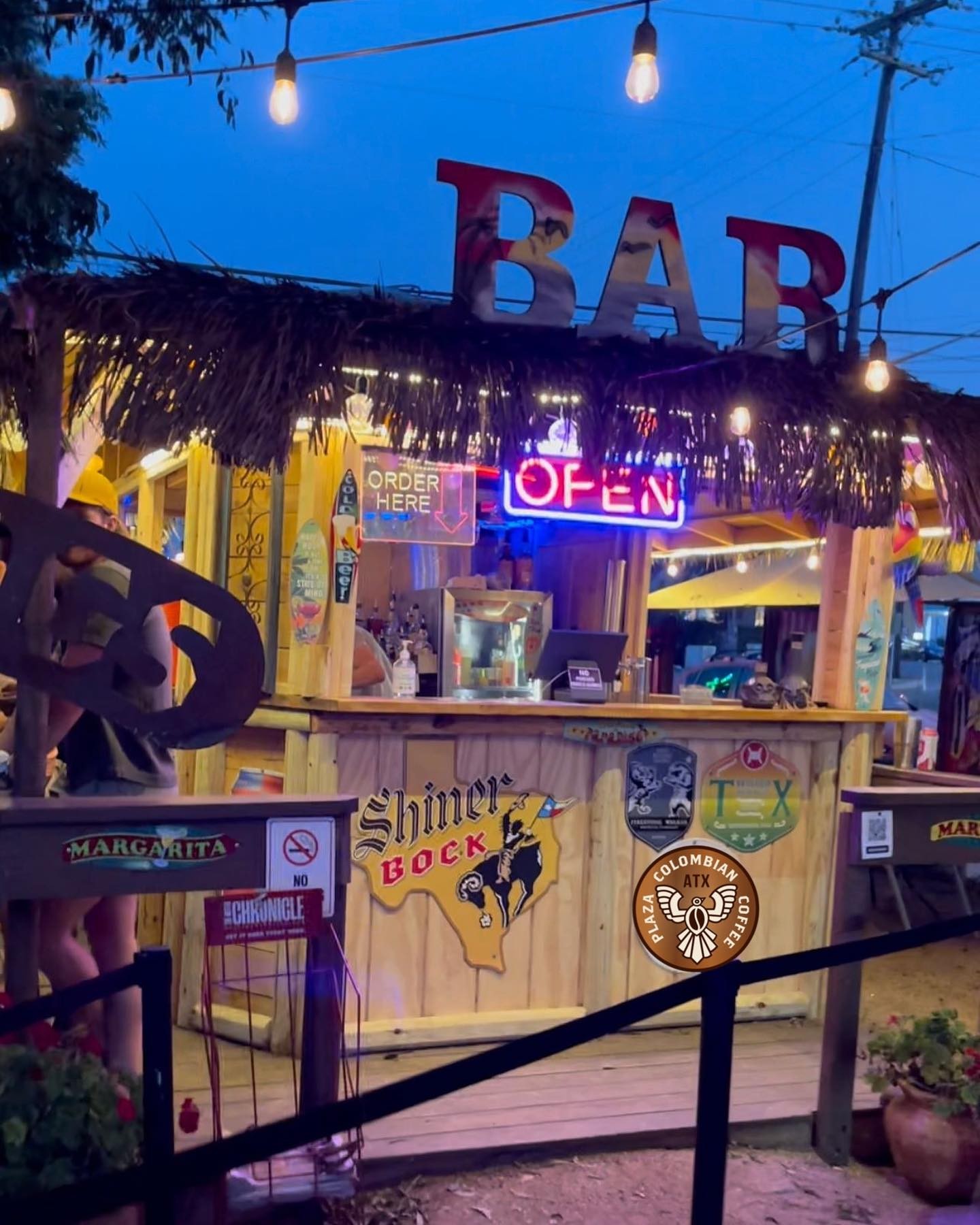 Thatch-roofed outdoor bar with "OPEN" neon sign and string lights at dusk.