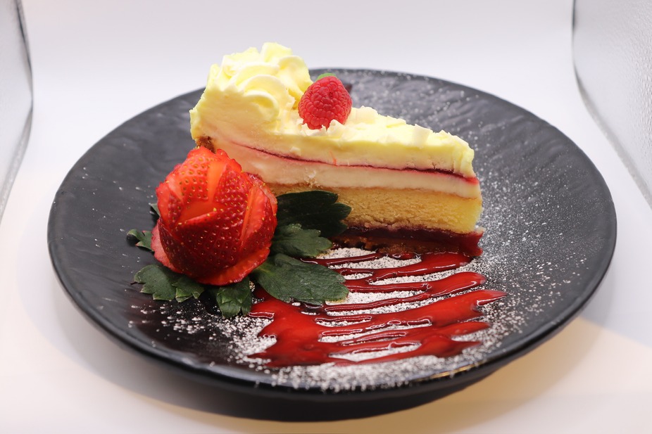 Slice of layered cake with raspberry, strawberry rose, syrup on black plate.