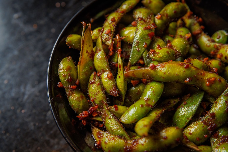 Green edamame pods covered in red seasoning in a black bowl.