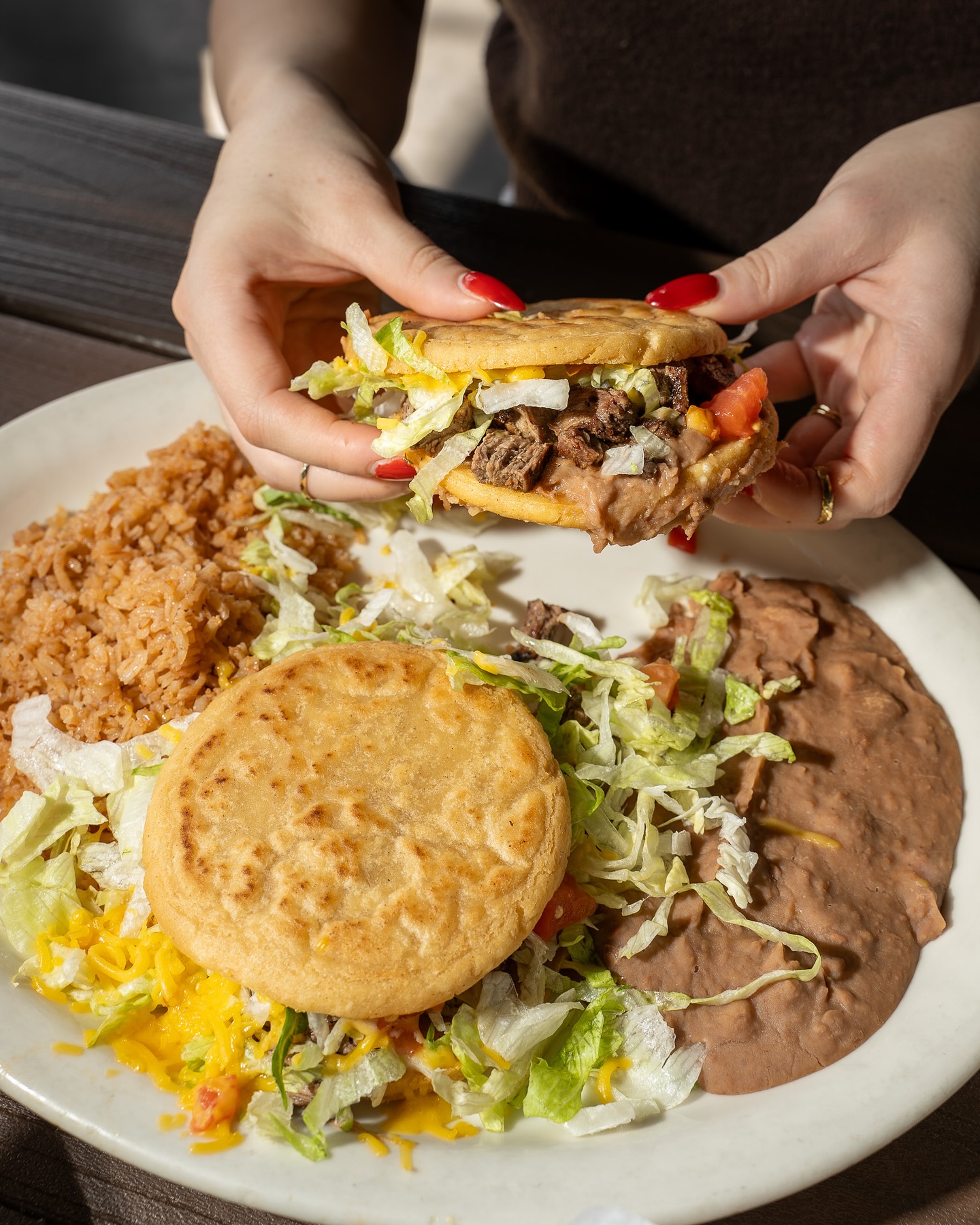 Hands holding a stuffed corn cake, with rice and beans on a plate.