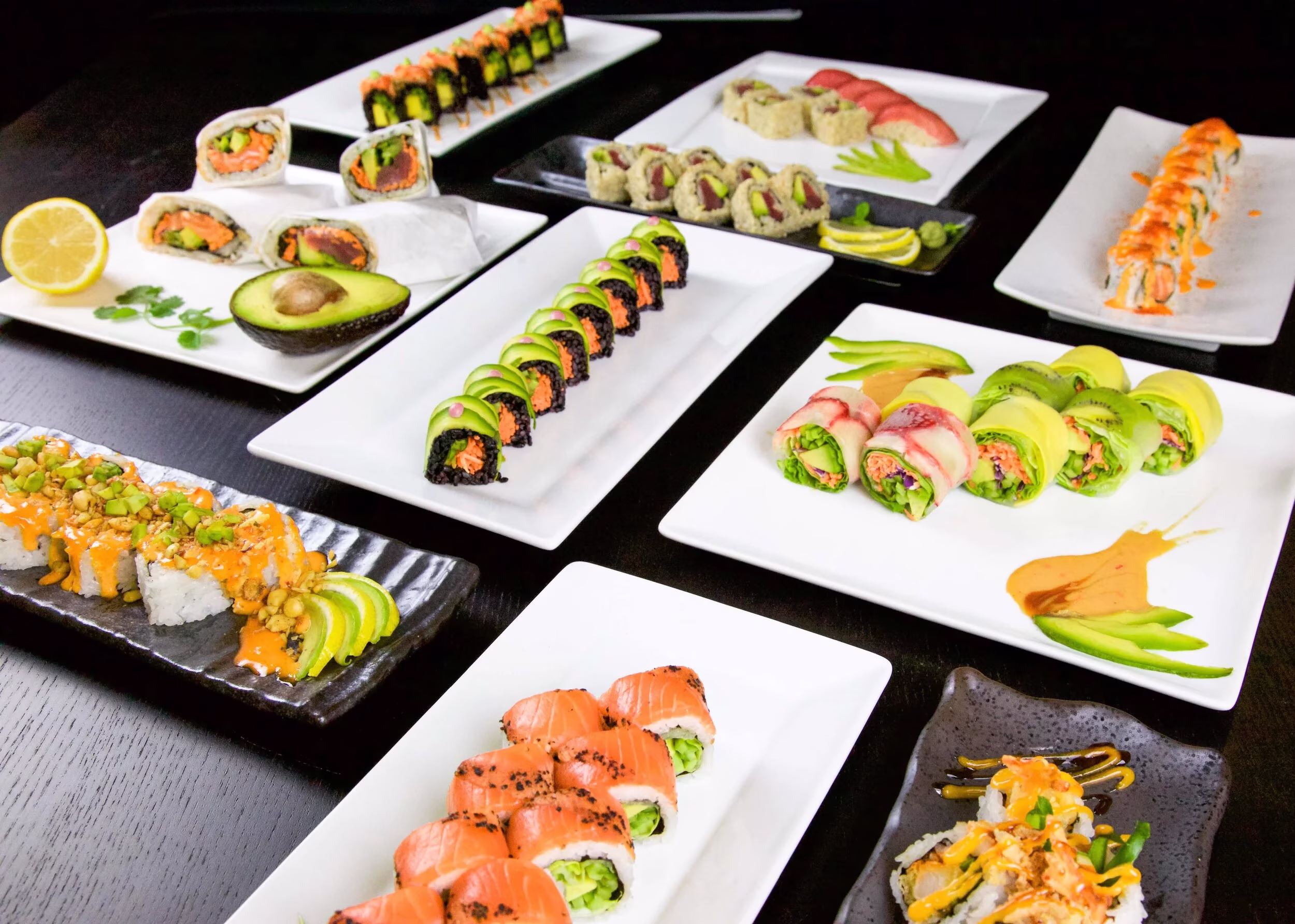 An assortment of different sushi rolls on multiple white and dark plates.