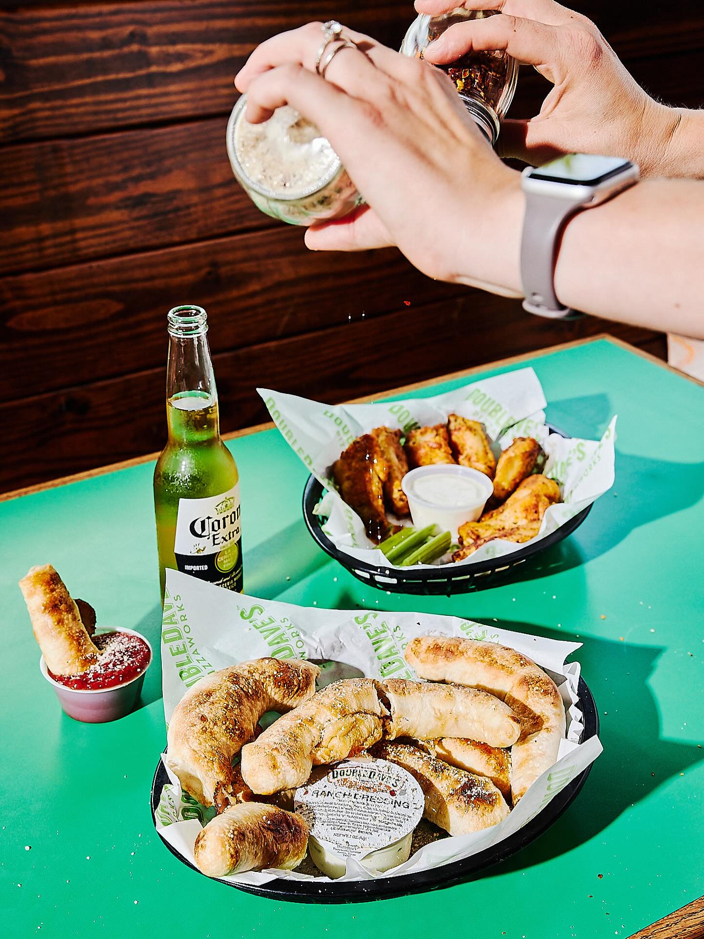 Hands seasoning a meal of breadsticks, chicken wings, and beer.