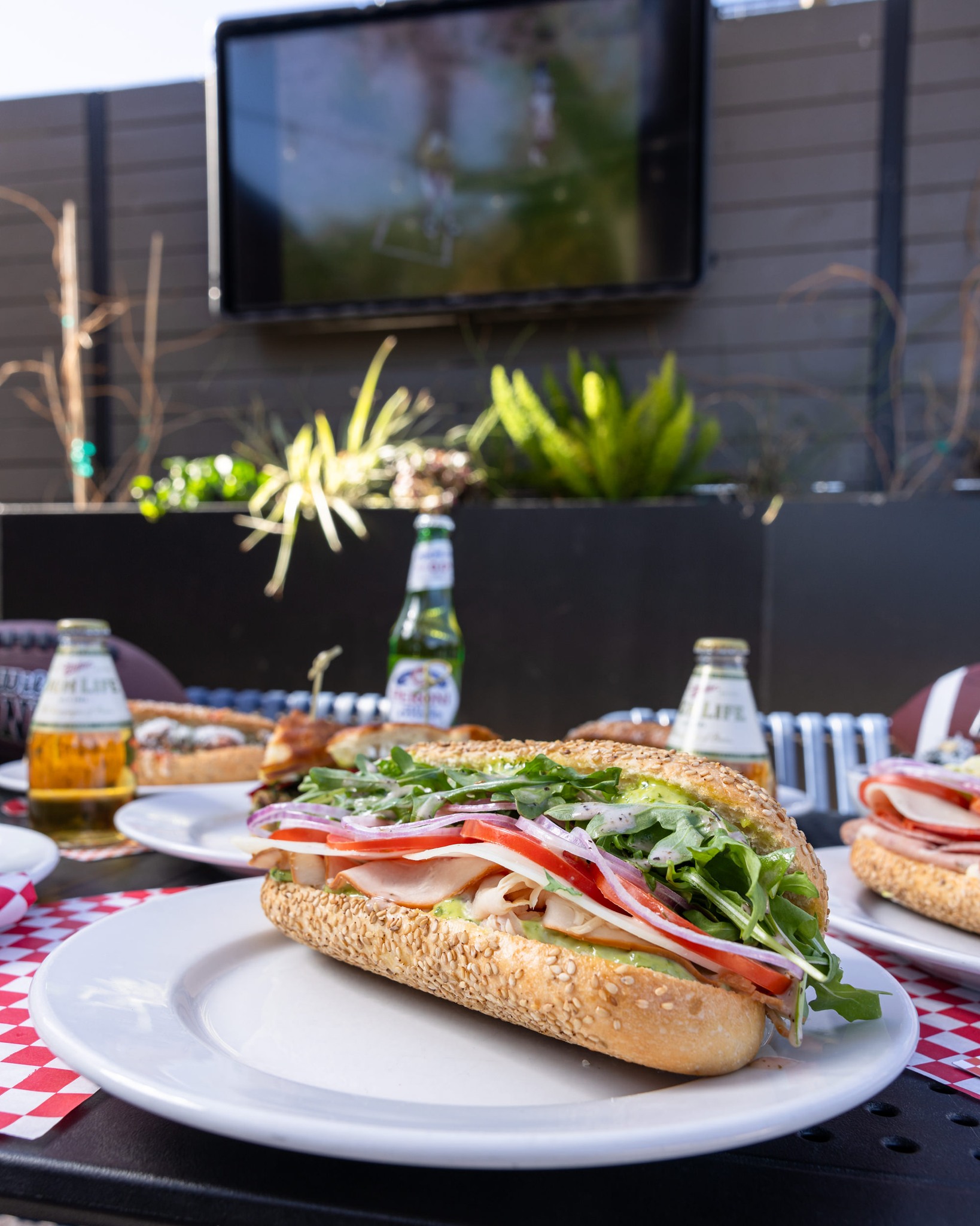 Sesame seed sub sandwich, beer bottles, and blurred outdoor TV.