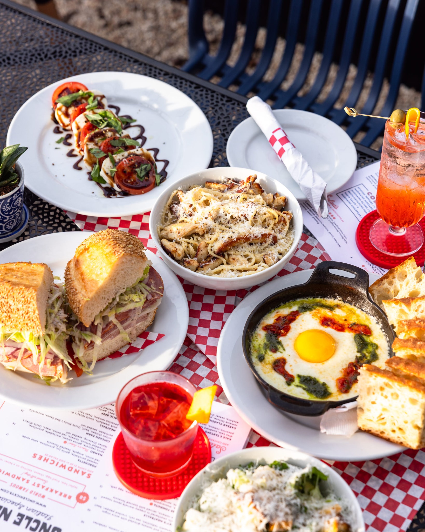 Outdoor table with Caprese salad, pasta, sandwich, baked egg, and cocktails.