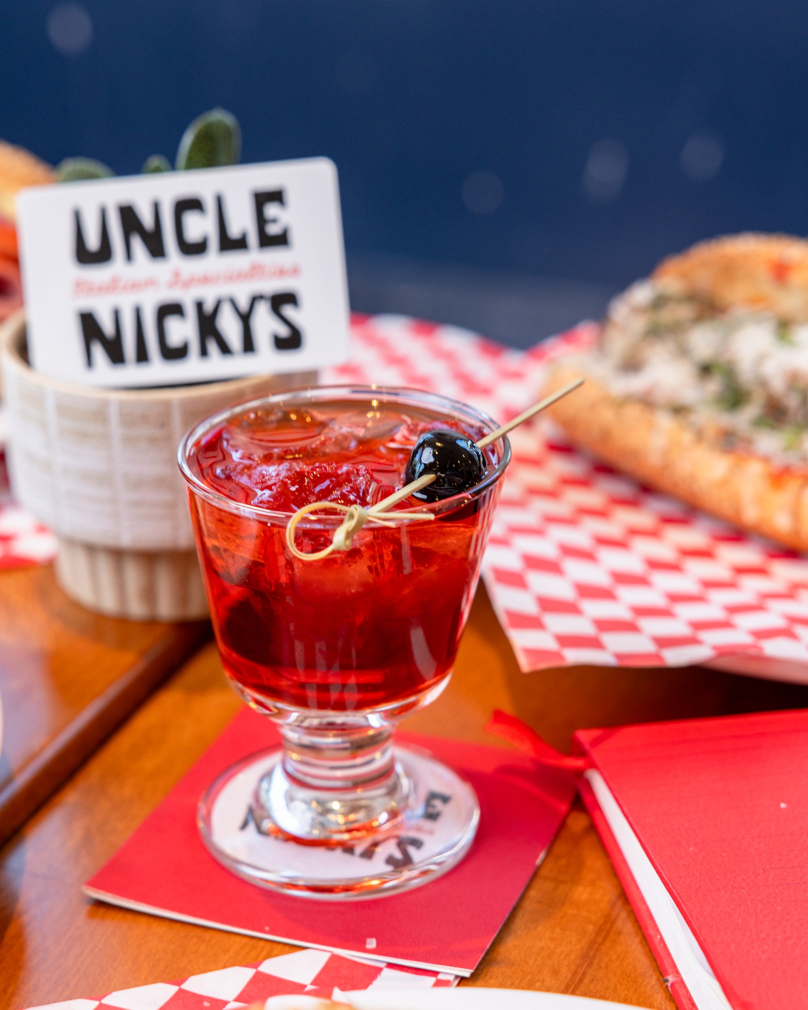 Red cocktail with cherry, Uncle Nicky's sign, and blurry sandwich.