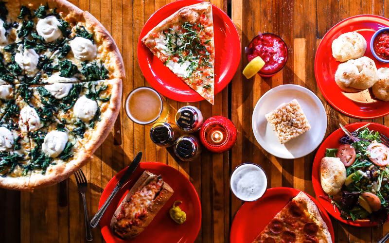 Pizzas, sub, salad, and drinks covering a rustic wooden table.