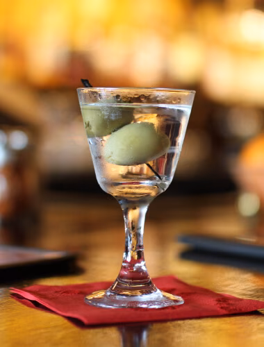 Stemmed glass with clear drink and two green olives on red napkin.